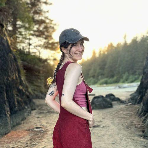 A white person with long brown hair in a single braid standing on a sandy beach between two large rocks. They are wearing a grey baseball cap, a pink tank top, red overalls, no shoes, and have a few tattoos on their arms and calves.
